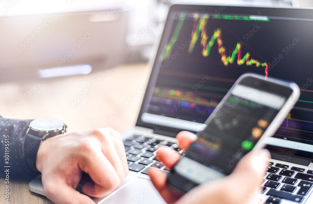 Man holding  smartphone in hand and working on laptop, financial bitcoin crypto graph data trade.  Business work on his computer at office desk, Typing keyboard detail.